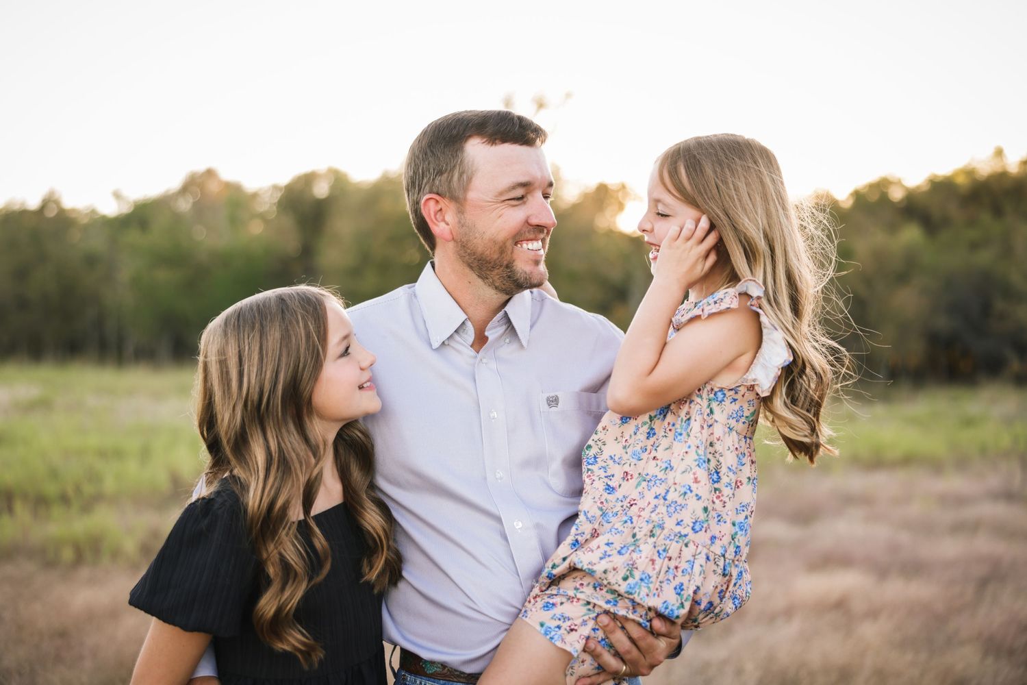 A father smiles joyfully holding his young daughter while his older daughter looks up at him in a sunny field.