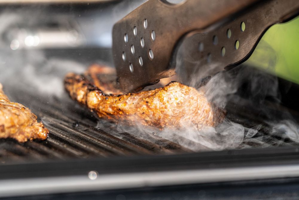 Smoky grilled chicken on a BBQ grill being flipped with metal tongs, with smoke rising from the hot grates.