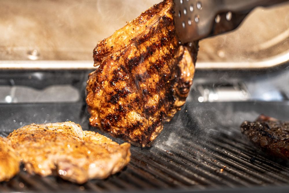 Juicy grilled pork chops cooking on a BBQ grill, being lifted with metal tongs showing perfect char marks.