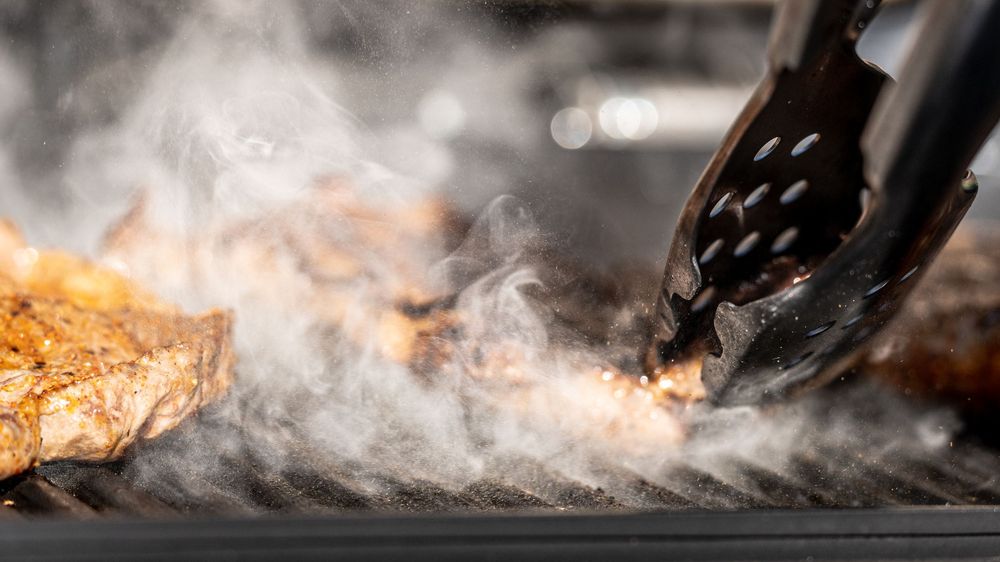 Tongs flipping juicy grilled meat on a smoky BBQ grill, perfect for outdoor cooking and grilling enthusiasts.