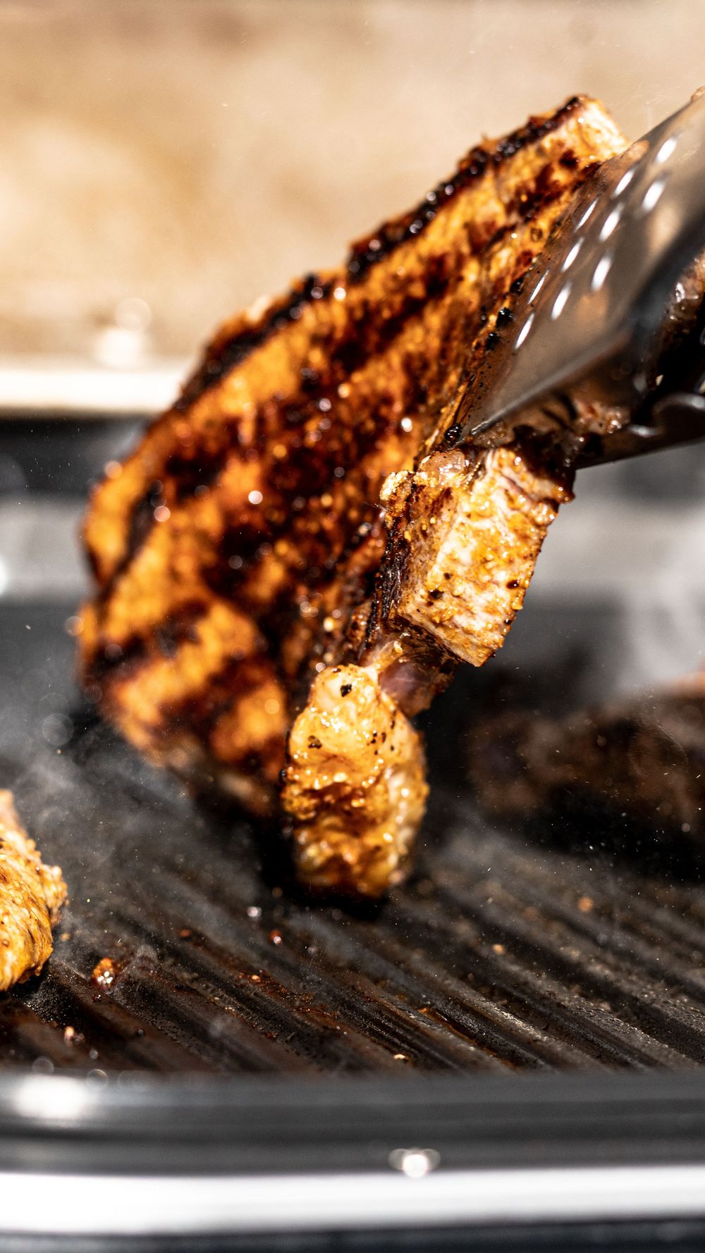 Grilled meat being lifted with tongs from a hot grill pan, showing charred marks and crispy texture.