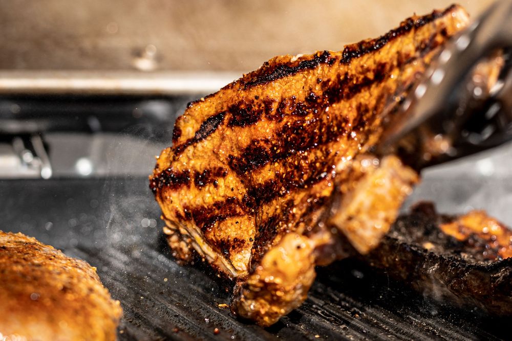Grilled meat with char marks being lifted by tongs on a hot grill, showing a perfectly caramelized crust.