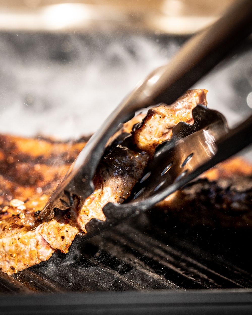 Tongs gripping smoky, charred grilled meat on a hot barbecue grill with golden-brown seasoning.