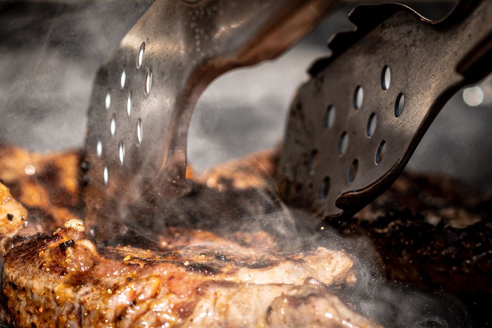 Tongs gripping a sizzling grilled steak with steam rising on a hot barbecue grill.
