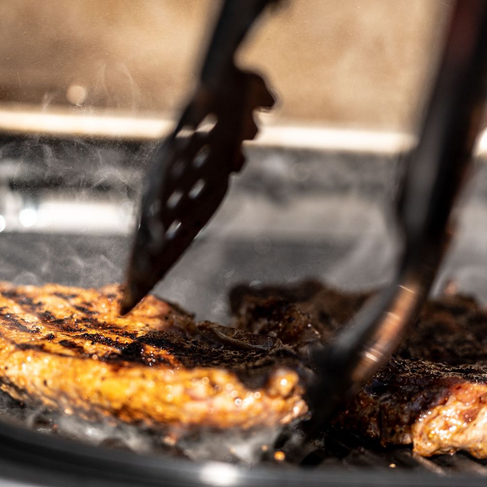 Close-up of grilled steak being handled with black tongs on a smoky BBQ grill with char marks.