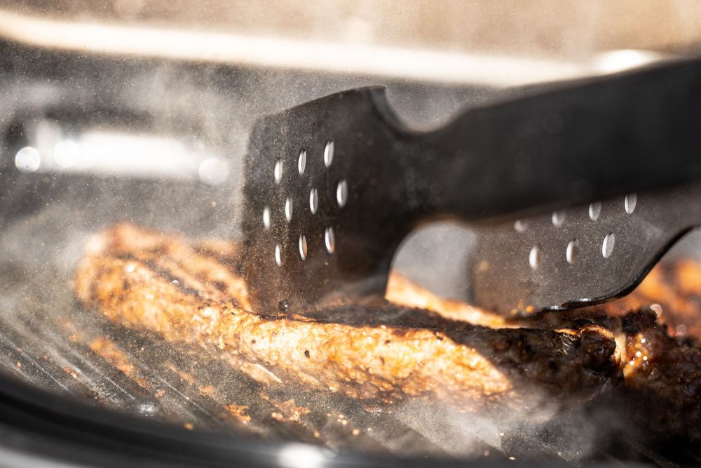 Close-up of a seasoned grilled steak being flipped with black spatulas on a smoking hot grill pan.
