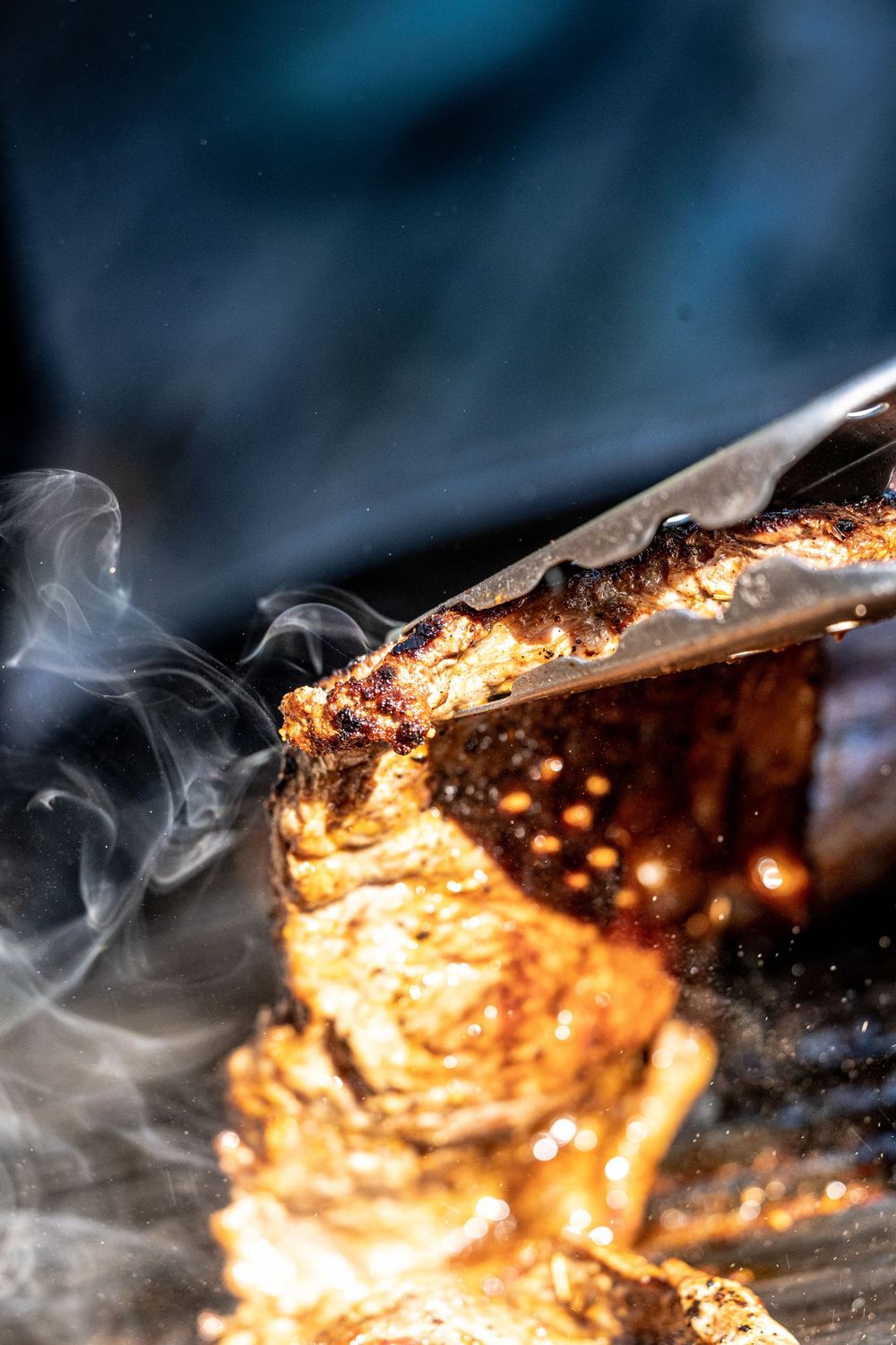 Close-up of sizzling grilled meat held by tongs with smoke rising, golden charred crust glowing in warm light.