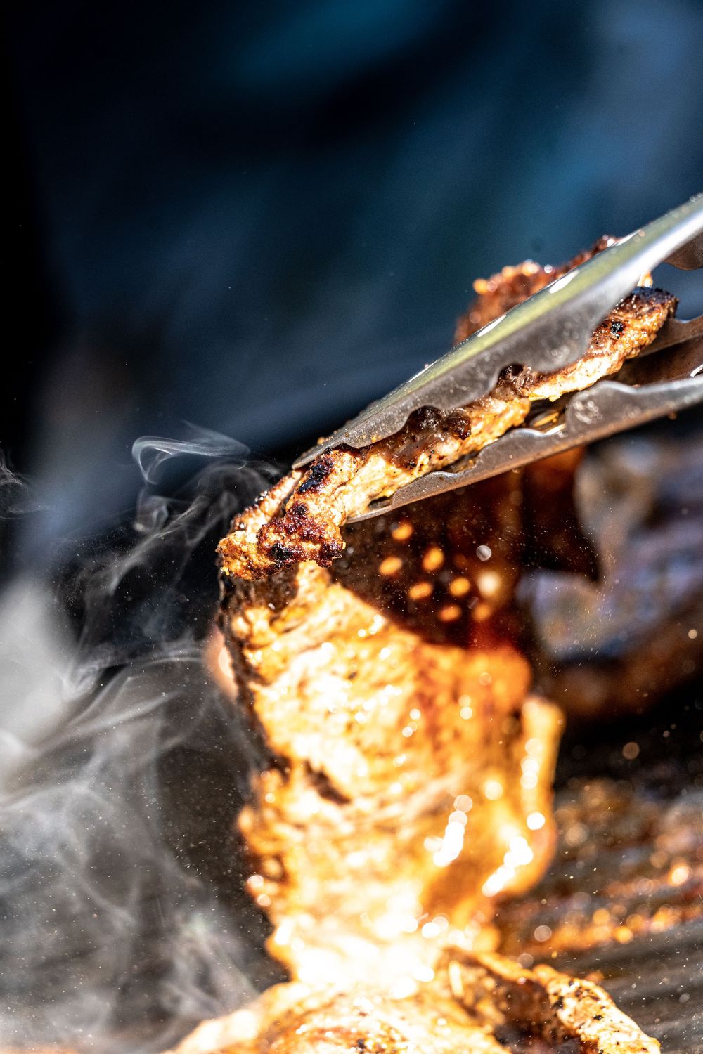 Juicy grilled meat held by tongs over open flames with smoke rising on a hot barbecue grill.