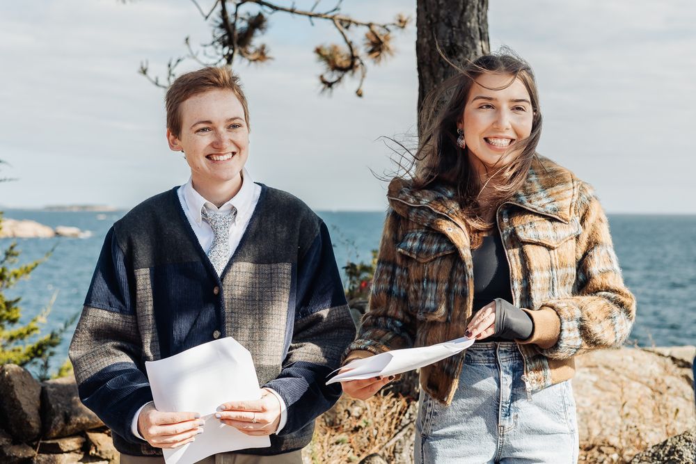 Two smiling young people holding papers outdoors near a rocky coastline on a sunny day.