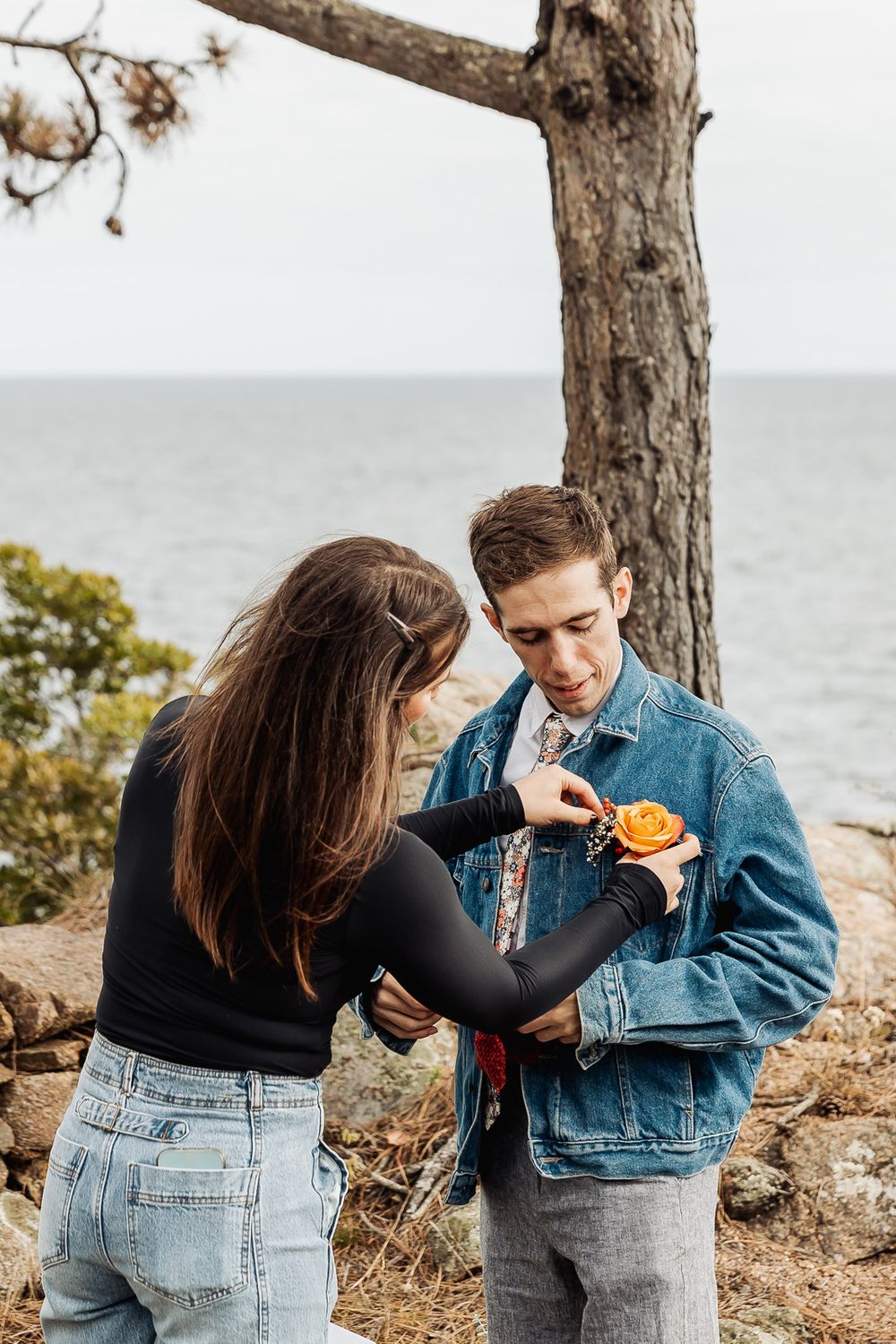 A woman pins an orange flower boutonniere on a mans denim jacket outdoors near the ocean.