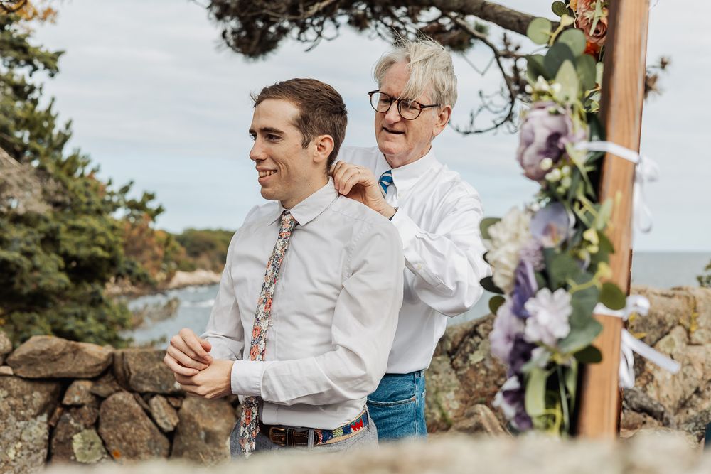 Older man adjusting younger man's collar outdoors at a seaside wedding ceremony with floral decorations.