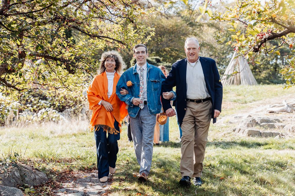 Three people walk outdoors on a sunny day, smiling and holding orange pumpkins on a scenic garden path.