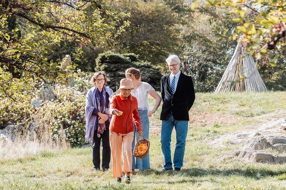 Four adults walking together on a sunny grassy path surrounded by autumn trees in a park setting.