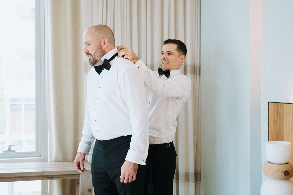 Groomsman adjusts bow tie of bald groom in white shirt before wedding ceremony.