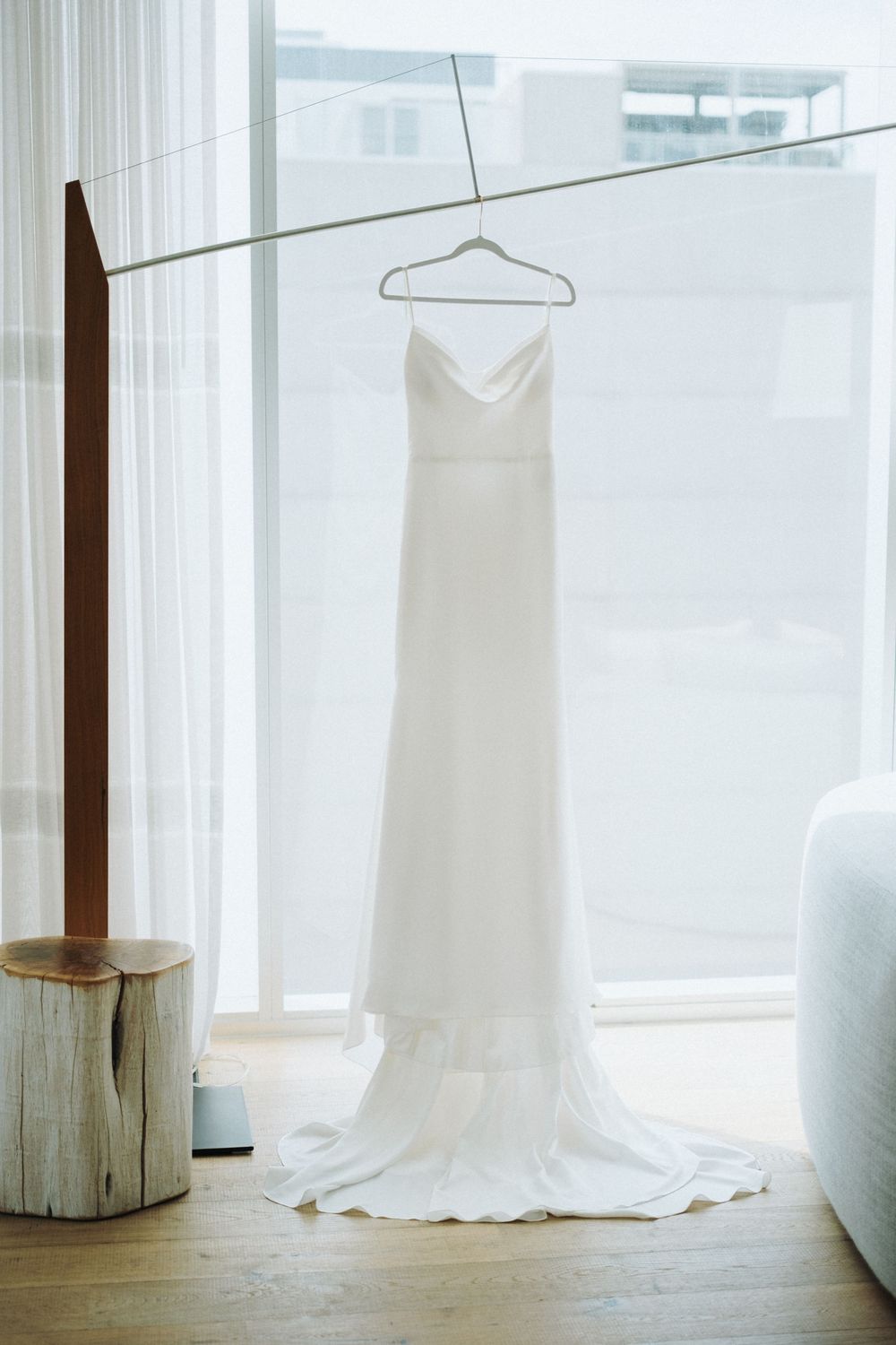 Elegant white bridal gown with a flowing train hanging on a hanger near a bright window in a modern room.