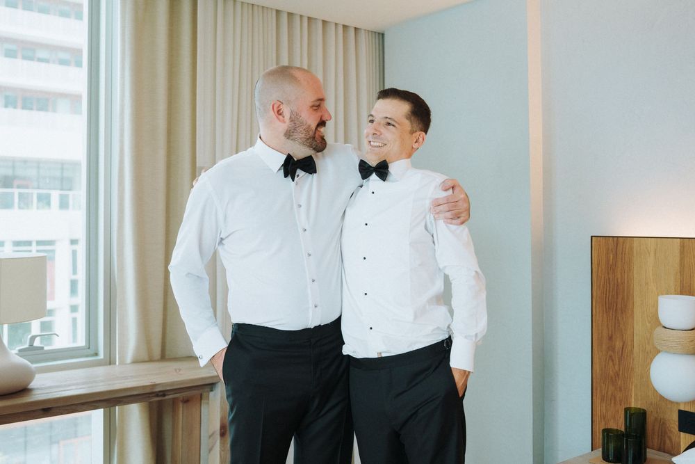 Two men in formal black and white tuxedo shirts smile together while getting ready for a wedding.