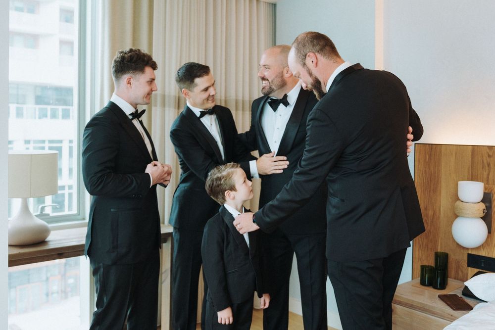 Groomsmen in black tuxedos chat with young ring bearer in hotel room before wedding.
