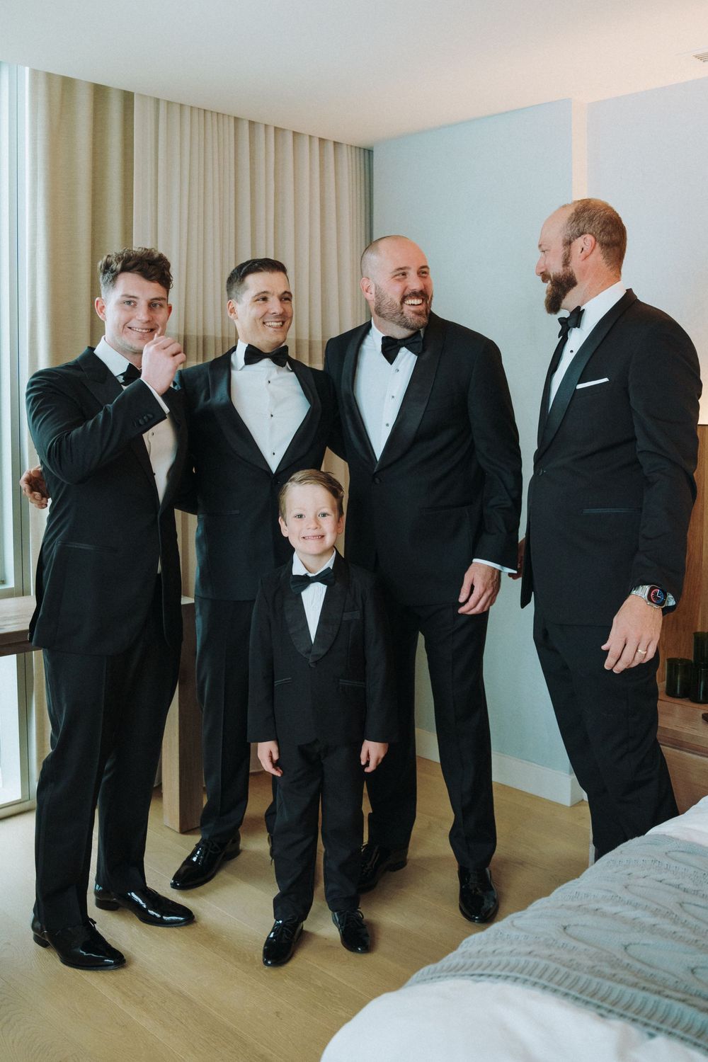 Four men and a young boy in matching black tuxedos laughing together in a hotel room before a wedding.