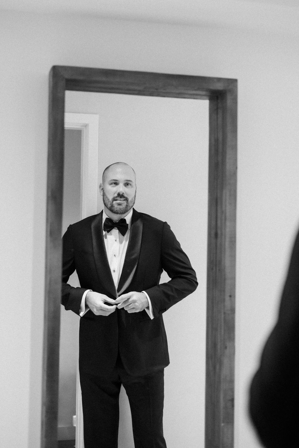 Dapper groom in black tuxedo and bow tie buttoning jacket while looking in full-length mirror.