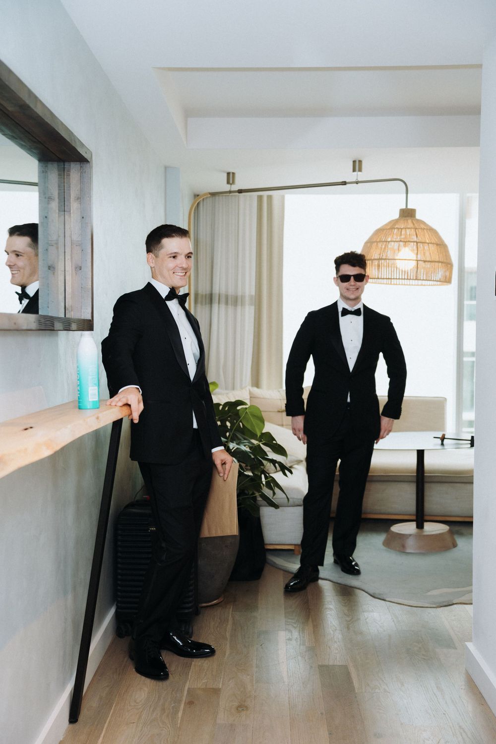 Two groomsmen in black tuxedos pose in a modern hotel room before the wedding ceremony.
