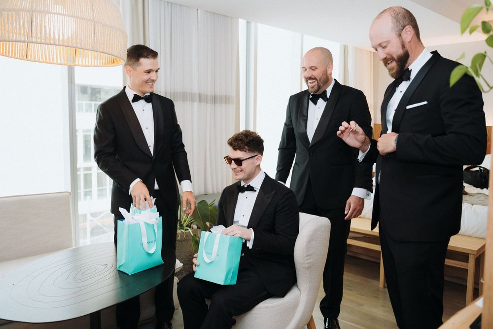Four groomsmen in black tuxedos celebrate with Tiffany gift bags in bright modern room before wedding ceremony.