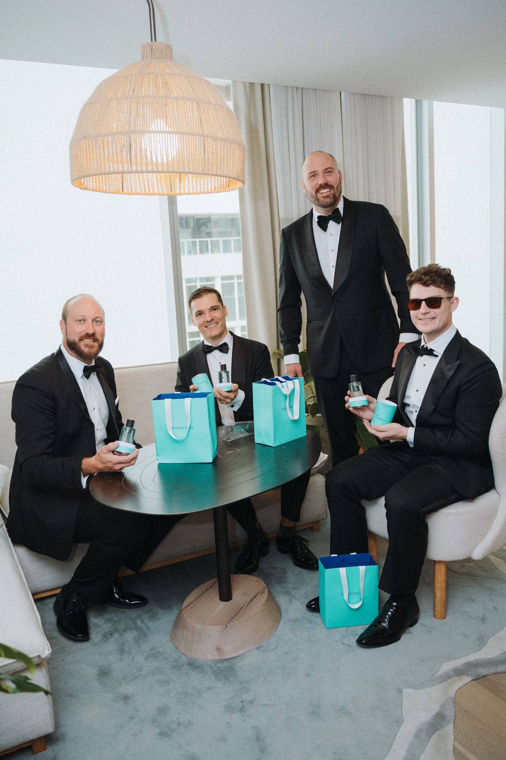Groom and groomsmen in black tuxedos celebrate with teal gift bags at hotel table.