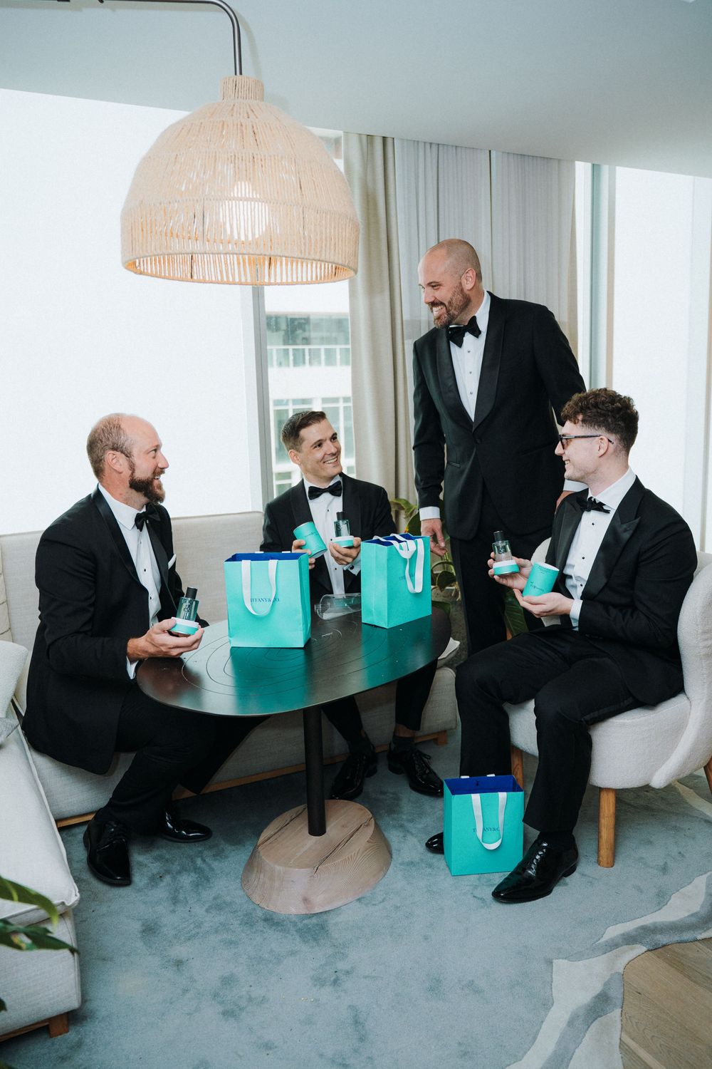 Groom and three groomsmen in black tuxedos laugh together with teal gift bags before wedding.