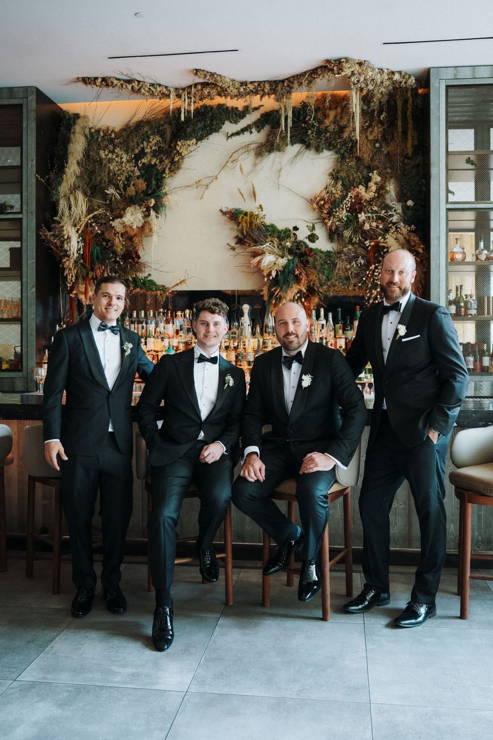 Four groomsmen in black tuxedos posing together at an elegant bar venue with botanical decor.