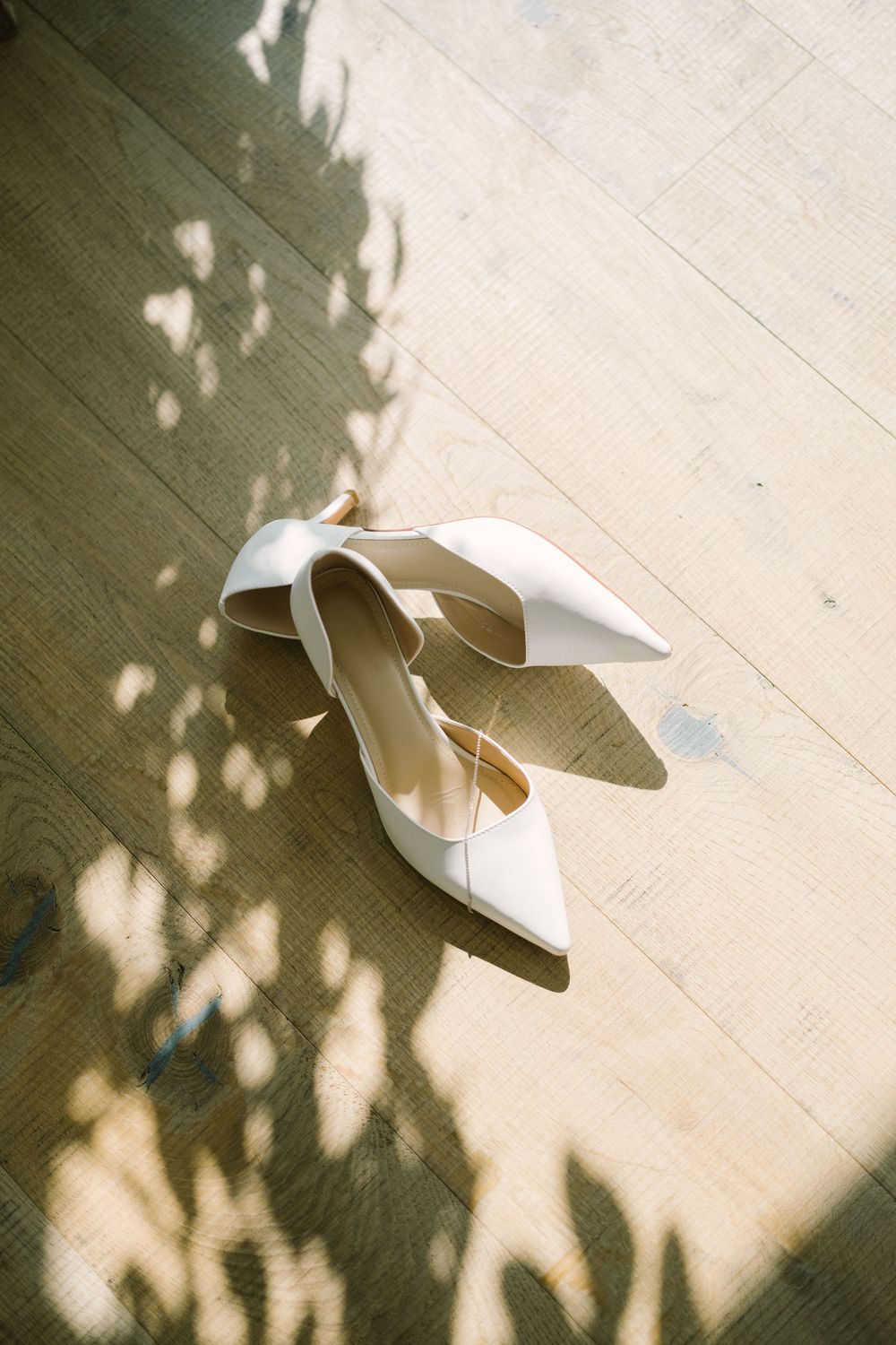 White satin bridal heels with pointed toes on a light wood floor with delicate leaf shadows.