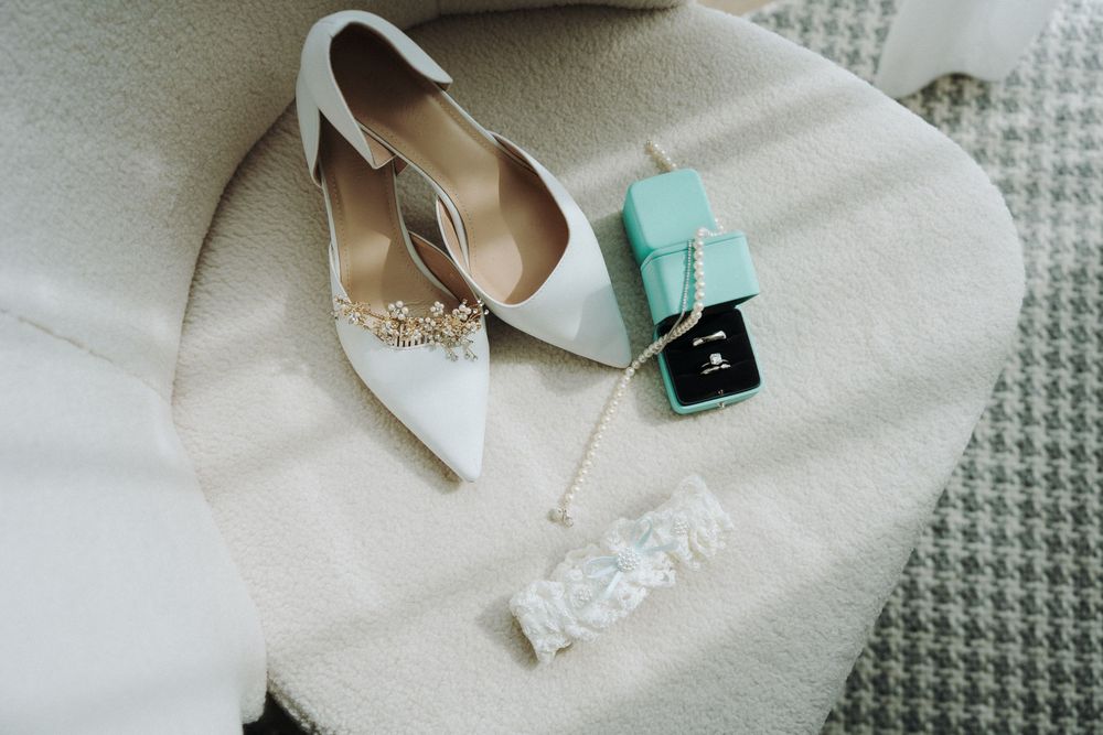 White bridal heels with crystal embellishment beside a Tiffany ring box, pearl necklace, and lace garter on a cream chair.