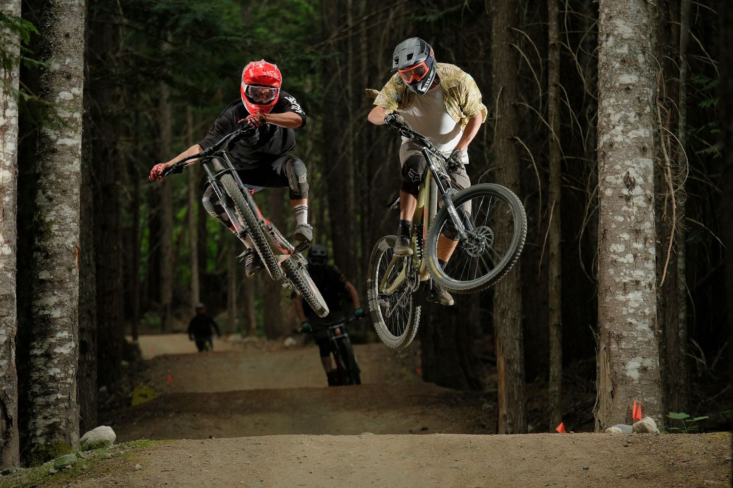 Two mountain bikers perform aerial jumps on a forest dirt trail wearing helmets.