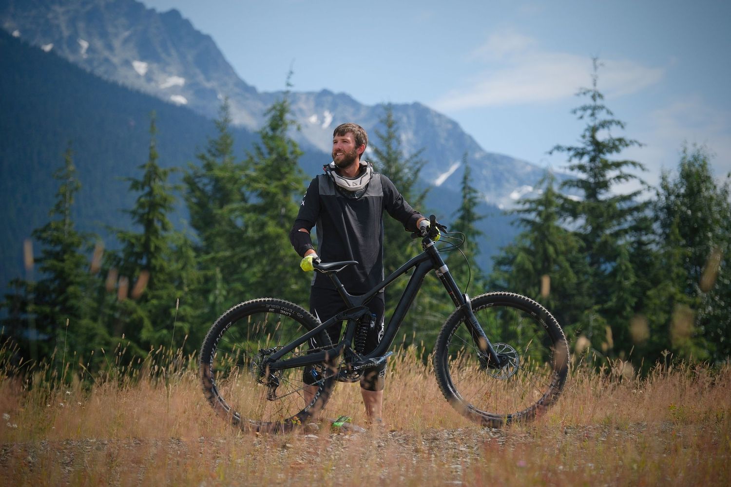 Man standing with black mountain bike in alpine meadow, evergreen trees and snow-capped mountains in background.