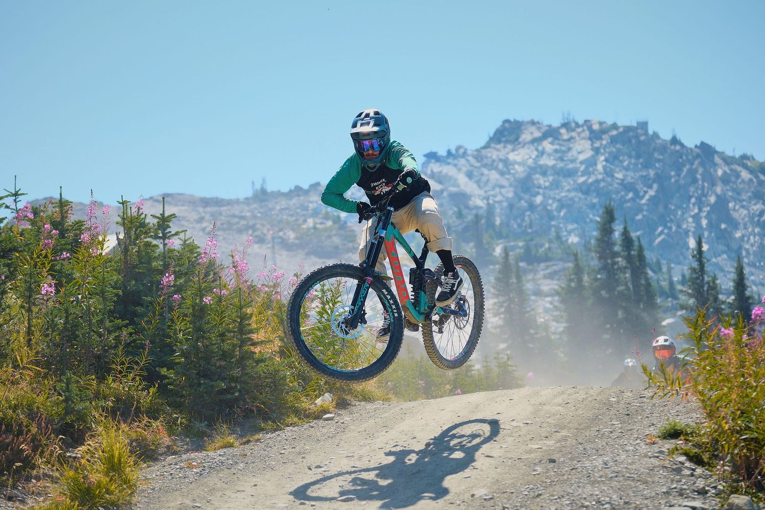 Mountain biker catching air on a dusty gravel trail with rocky peaks and wildflowers in the background.