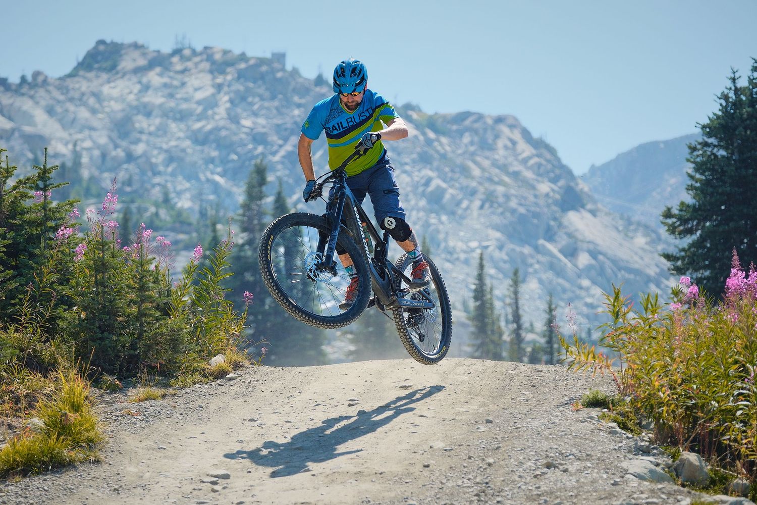 Mountain biker performing a jump trick on a dirt trail with rocky peaks and pine trees in the background on a sunny day.