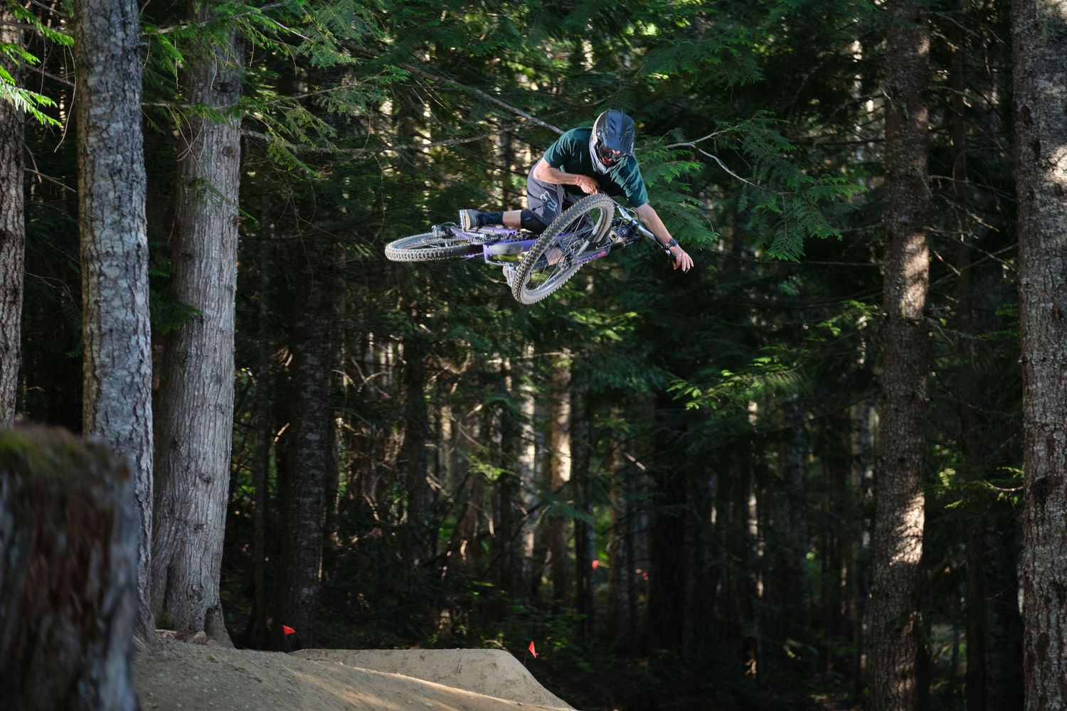 Mountain biker performs a high aerial trick jump in a dense forest bike park with tall trees in the background.
