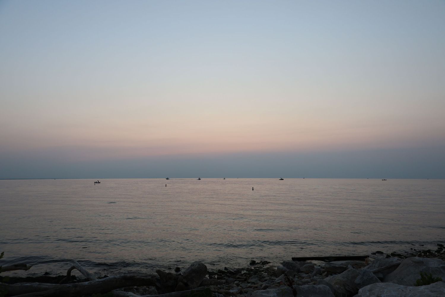 Serene dusk seascape with calm water, distant boats, and a soft pink and blue gradient sunset sky.