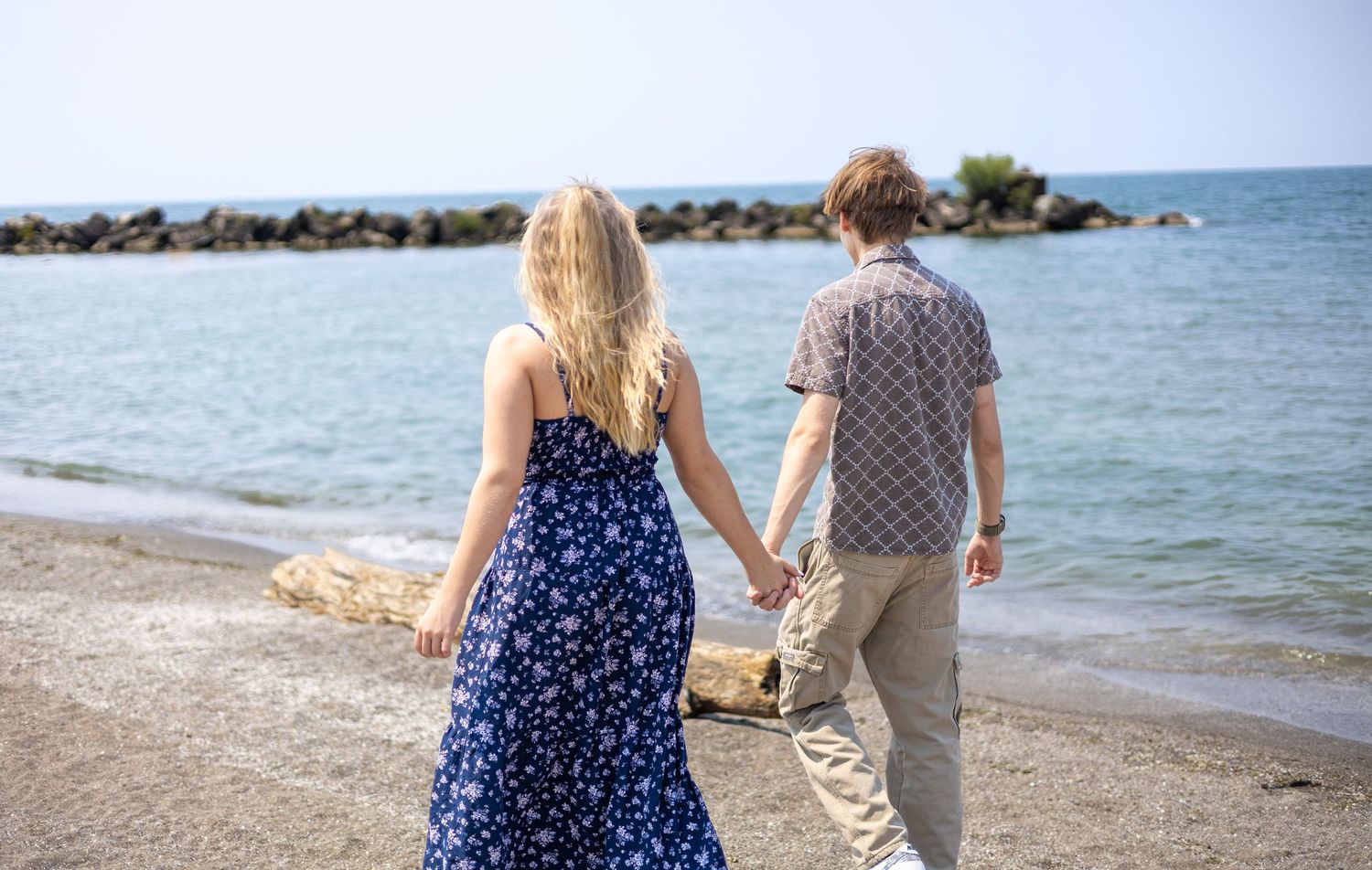 Young couple holding hands walking along a rocky shoreline beach on a clear sunny day.