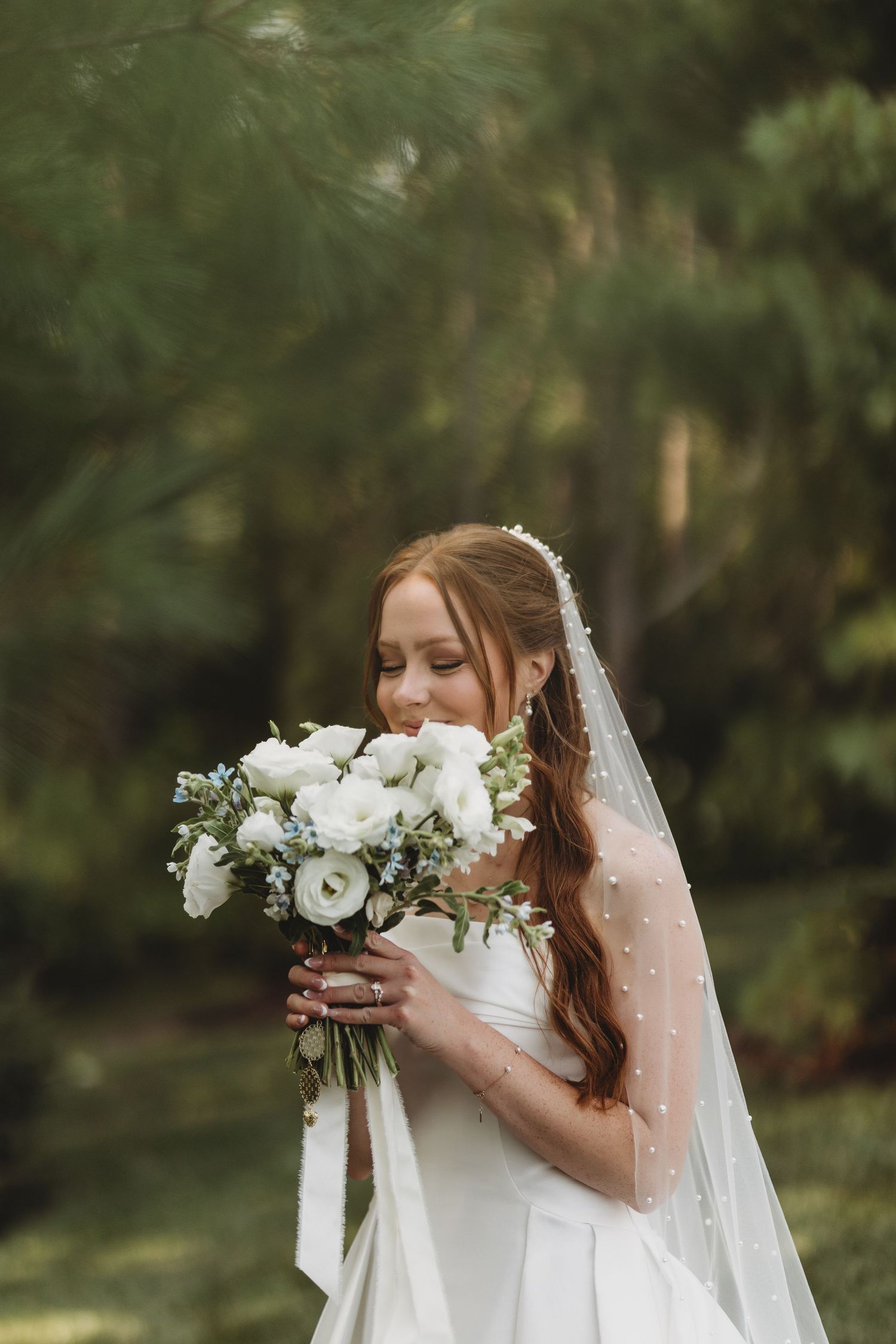 A bride in a white gown and pearl veil smells her white floral bouquet outdoors among lush green trees.