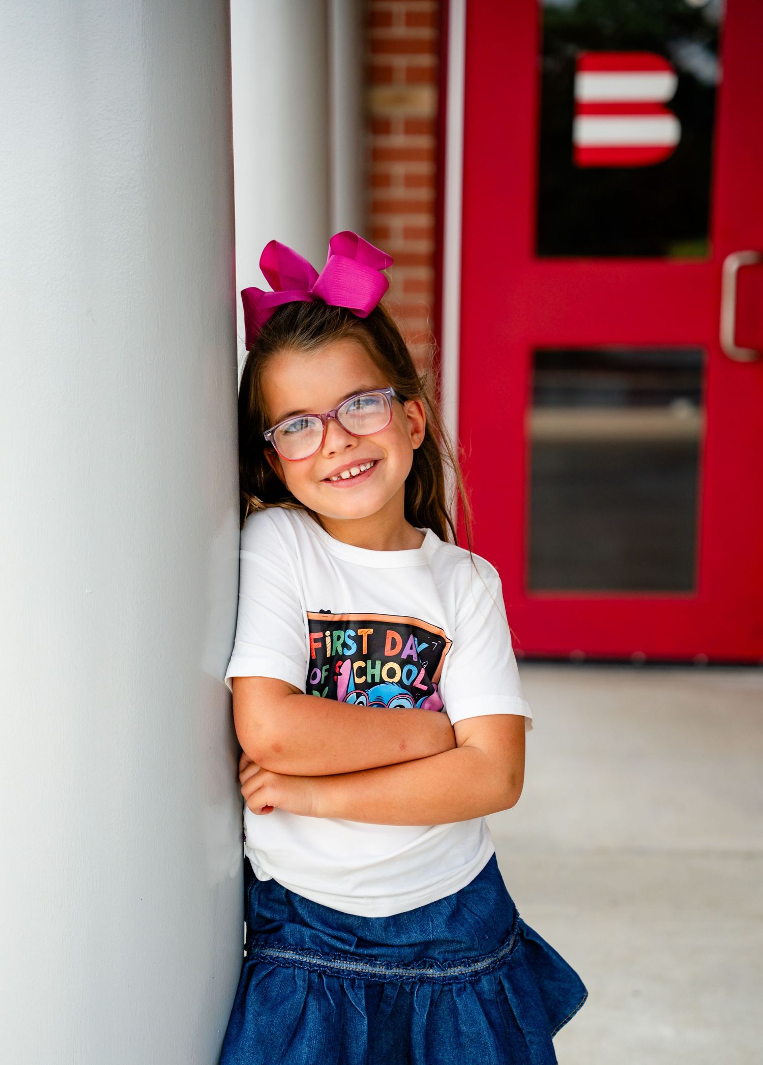 Smiling young girl in glasses and pink bow leans against pillar on first day of school.