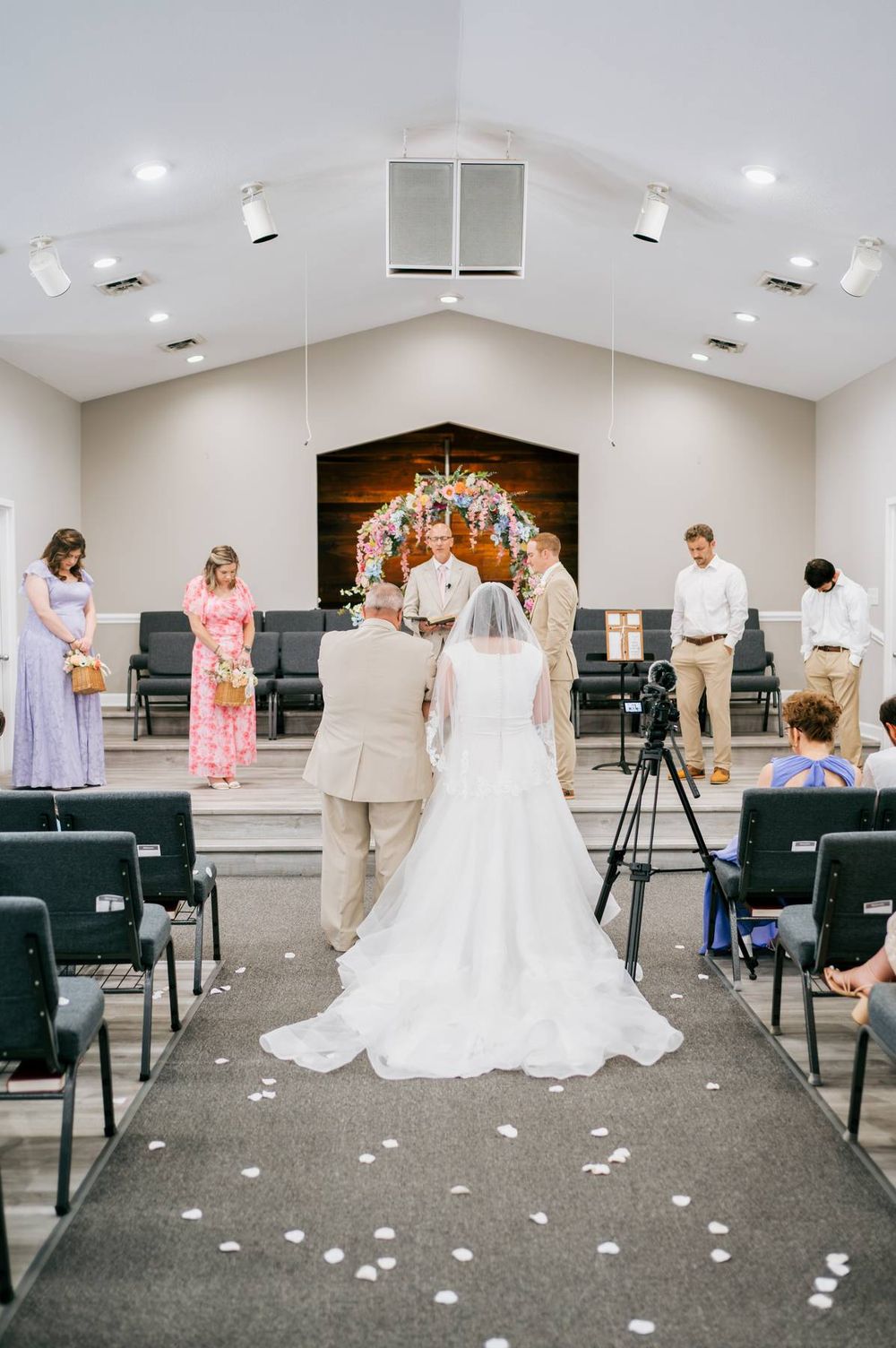 Bride walks down church aisle with escort toward colorful floral arch at indoor wedding ceremony, wedding photographer Asheboro NC