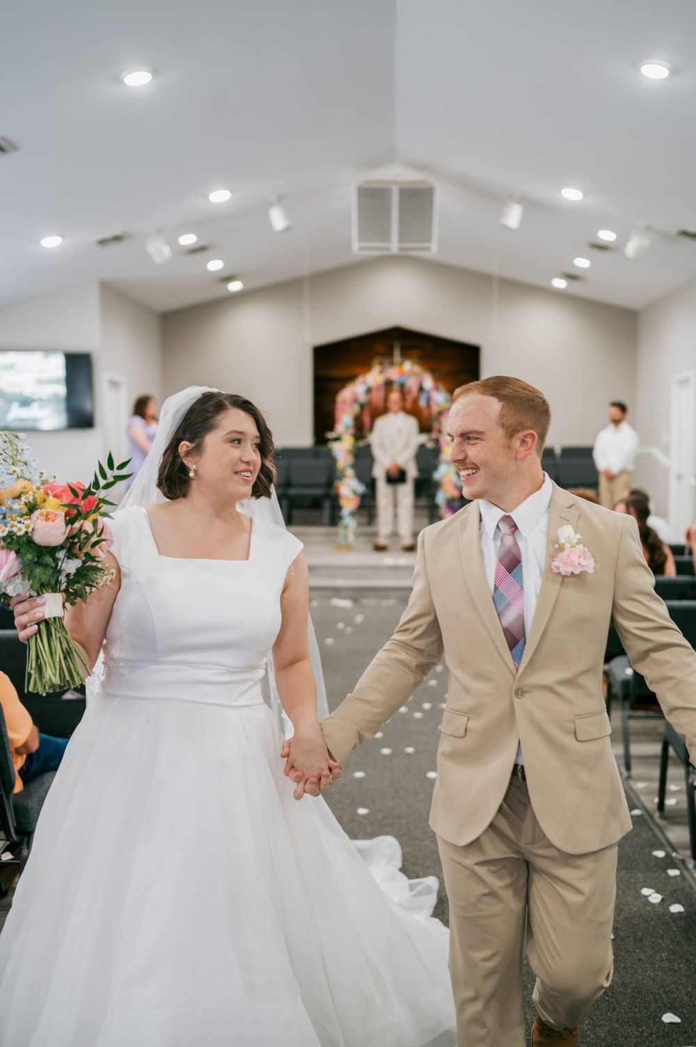 Smiling newlyweds walk hand in hand down church aisle after wedding ceremony with colorful bouquet, wedding photographer North Carolina