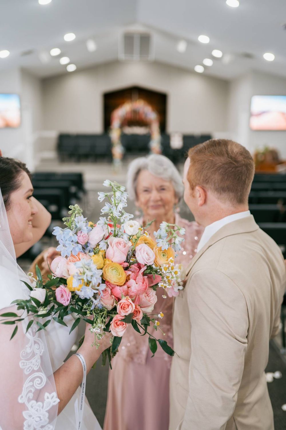 Elderly woman presents colorful pastel bridal bouquet to bride and groom at church wedding ceremony, wedding photographer Greensboro NC