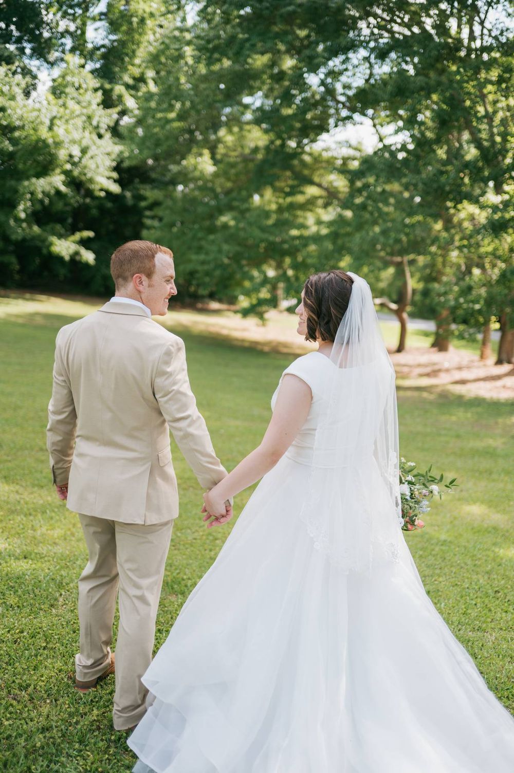 Bride and groom walk hand in hand through lush green park in wedding attire from behind, wedding photographer High Point NC