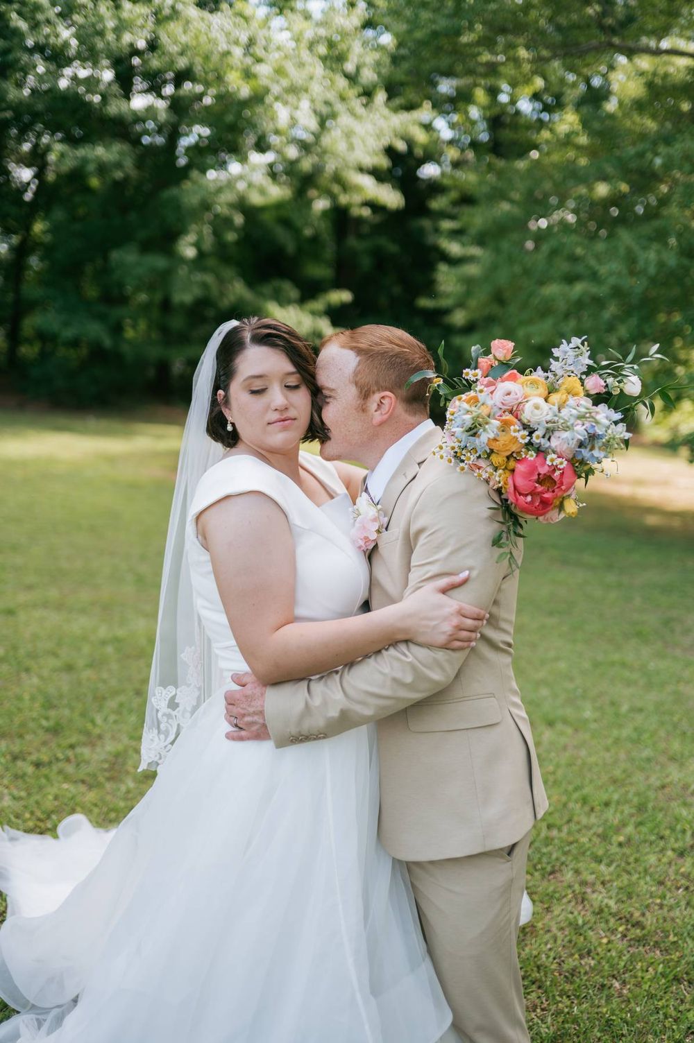 Groom in tan suit embraces bride while holding colorful bouquet of peonies outdoors in green park, wedding photographer Lexington NC