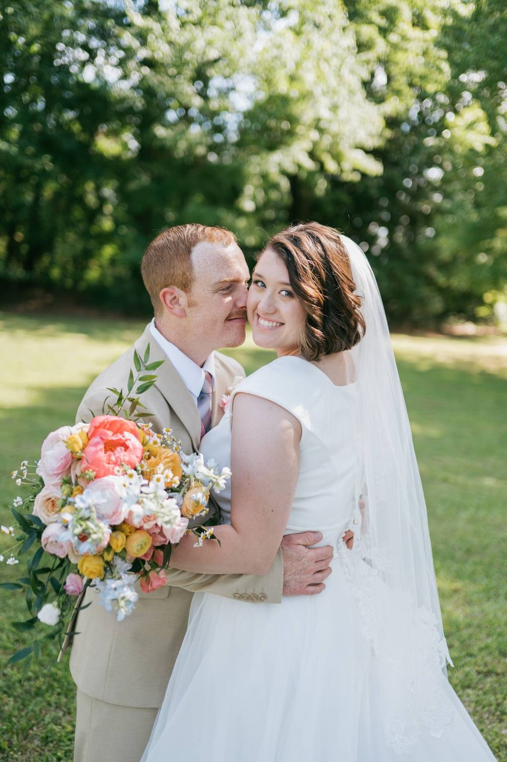 Groom kisses brides cheek as she smiles holding vibrant peony bouquet in sunny outdoor setting, wedding photographer NC