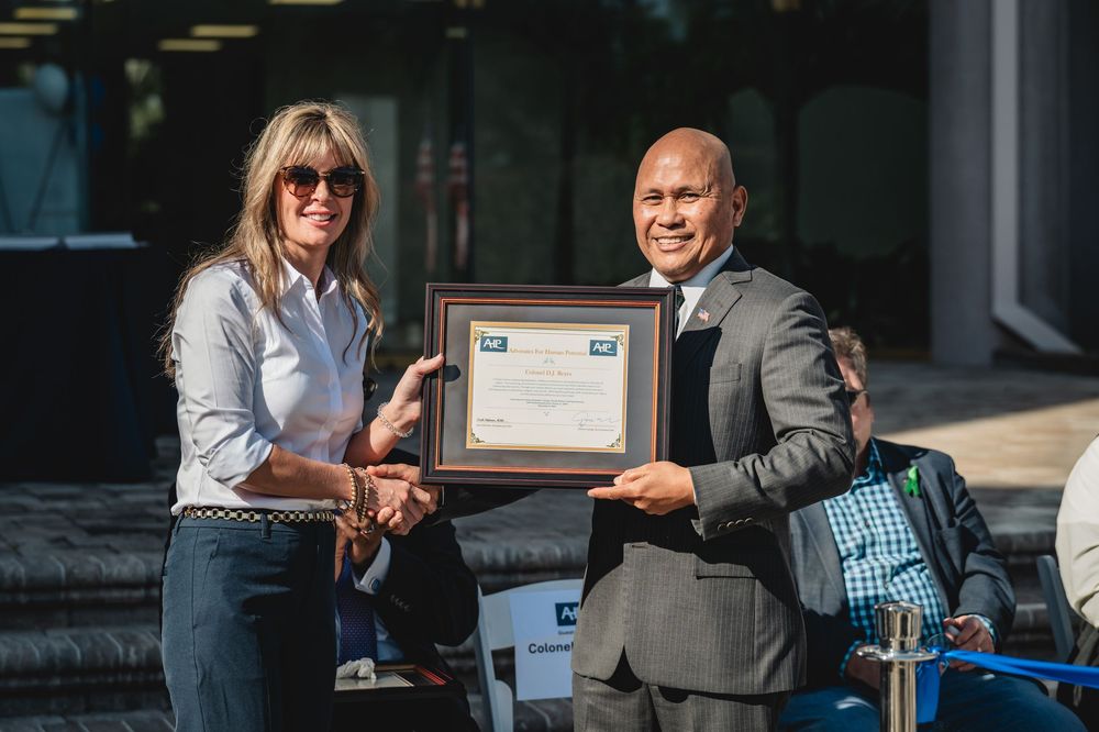 Two professionals exchange a framed certificate award outdoors at a formal recognition ceremony.