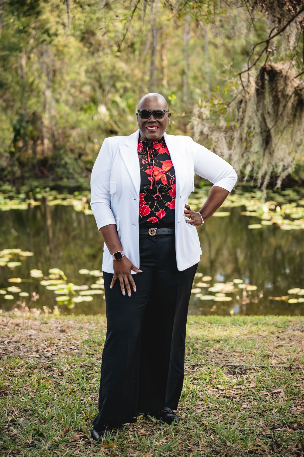 Woman in white blazer and floral top smiling outdoors near a pond with lily pads and lush greenery.
