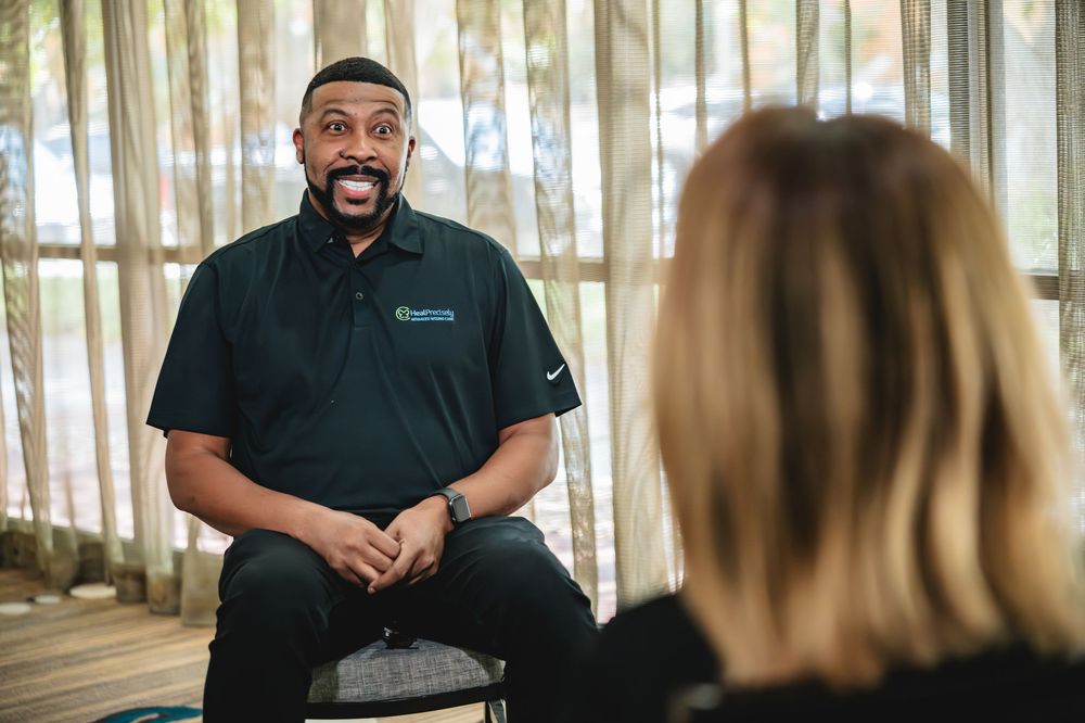 A smiling man in a dark polo shirt speaks animatedly to a person during an indoor meeting.