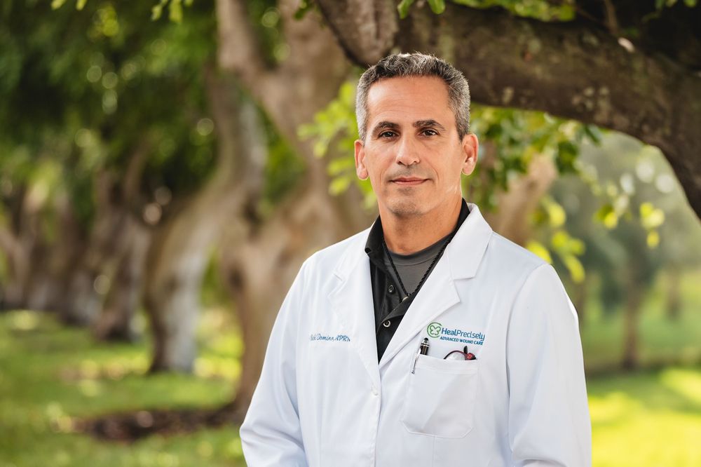 Male doctor in white lab coat standing outdoors under large trees in a professional portrait photo.