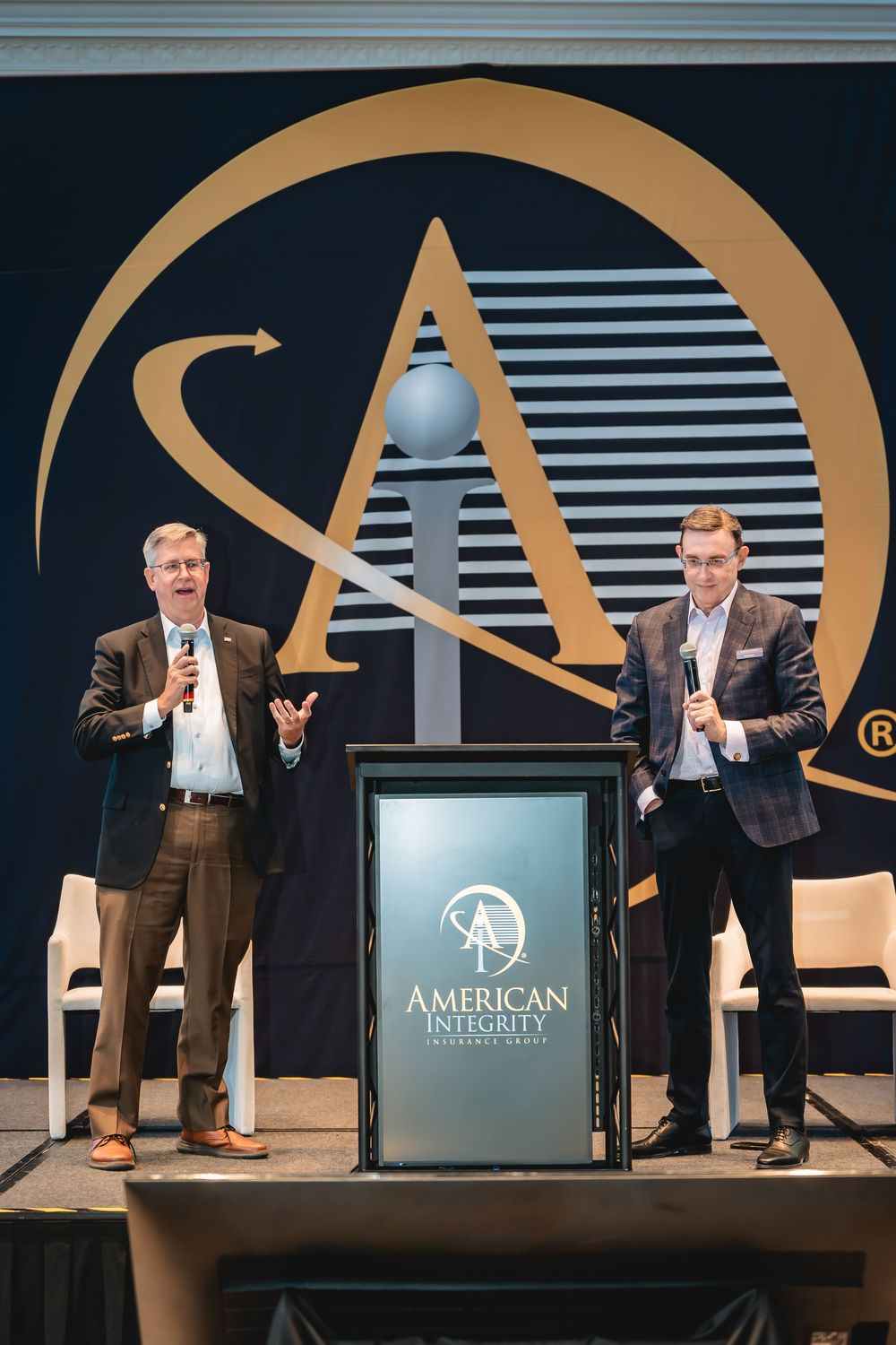 Two men speaking on stage at American Integrity Insurance Company event with branded podium and backdrop.